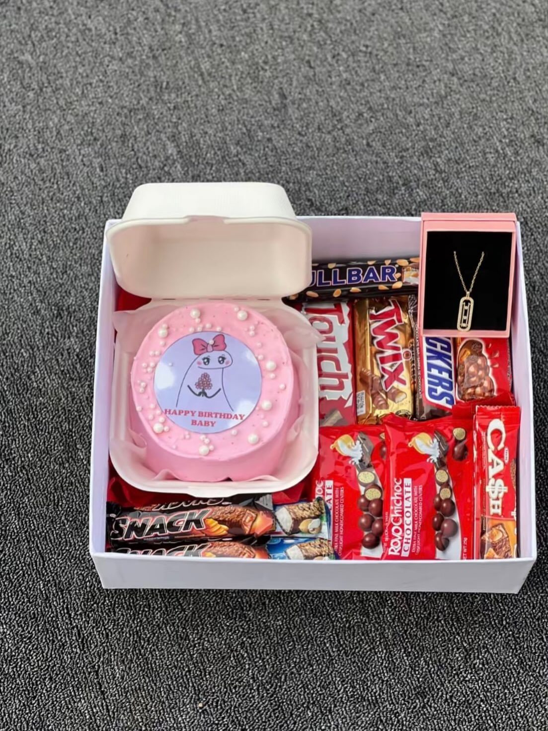 Pink birthday cake and assorted chocolates with a necklace in a white gift box.