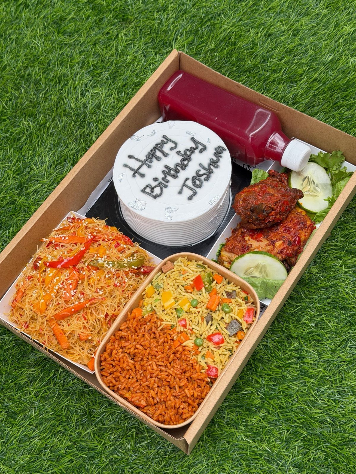 Birthday gift box with a small cake, red juice, chicken, and rice on grass.