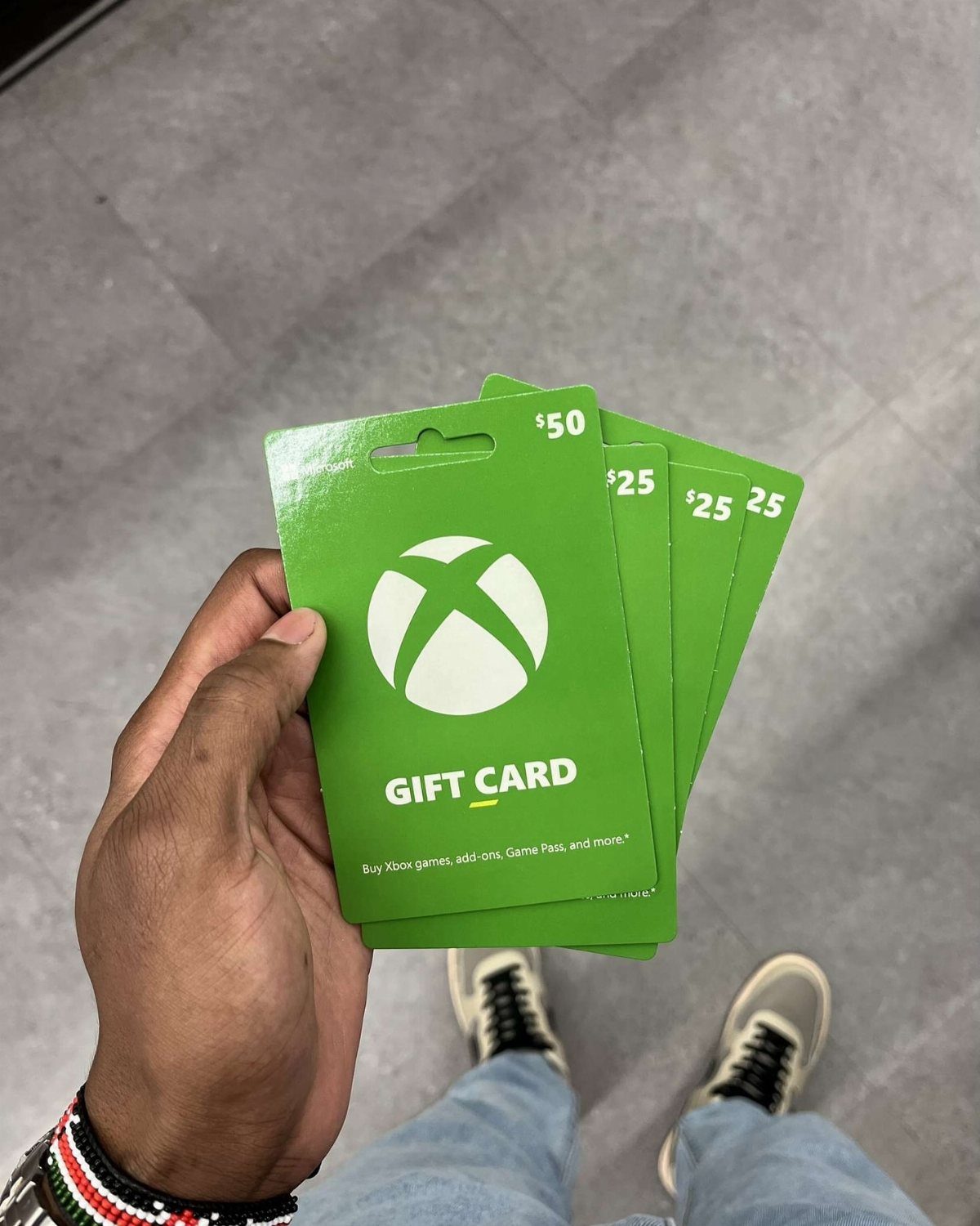 Hand holding four green Xbox gift cards over a grey tiled floor.