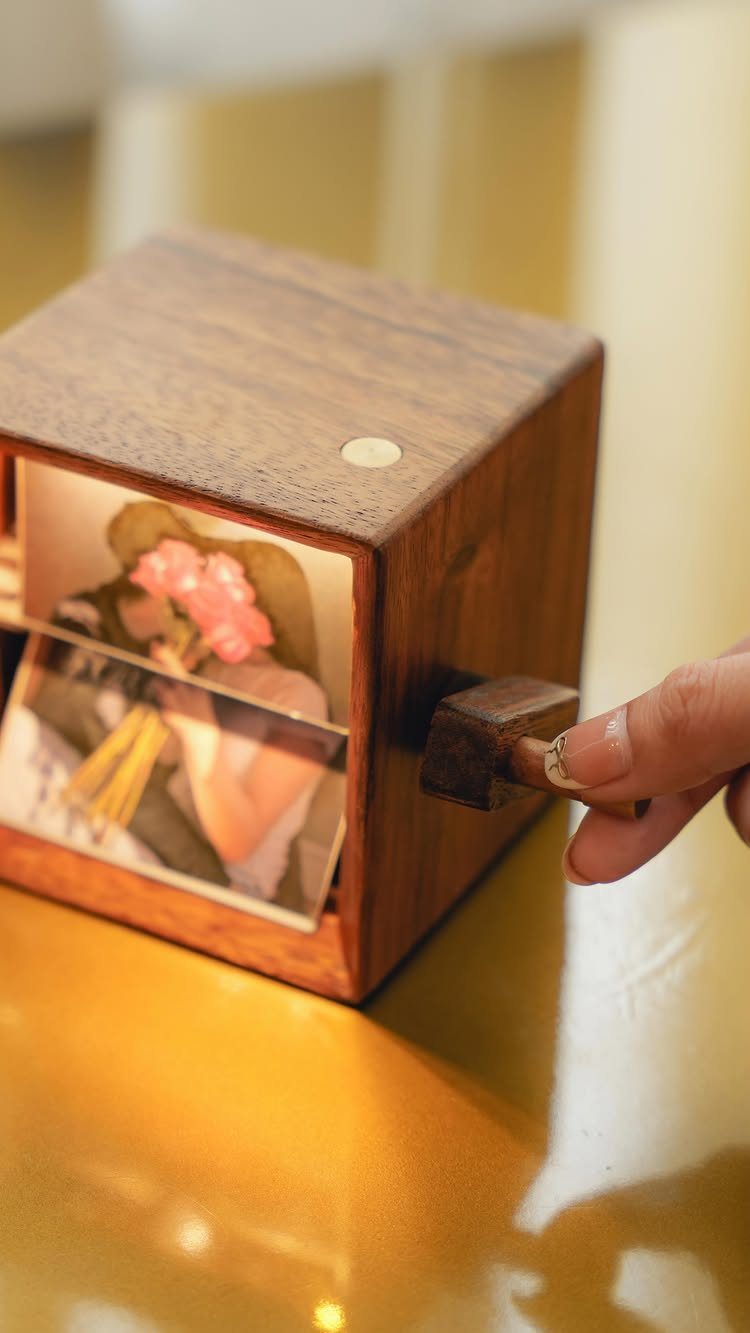 Wooden cubic music box with a photo window and a hand turning the crank.