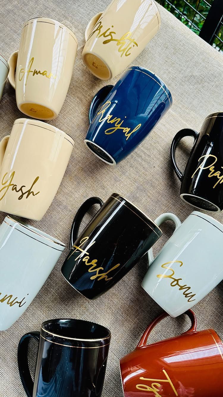 Various colorful ceramic mugs with personalized names written in gold cursive script.