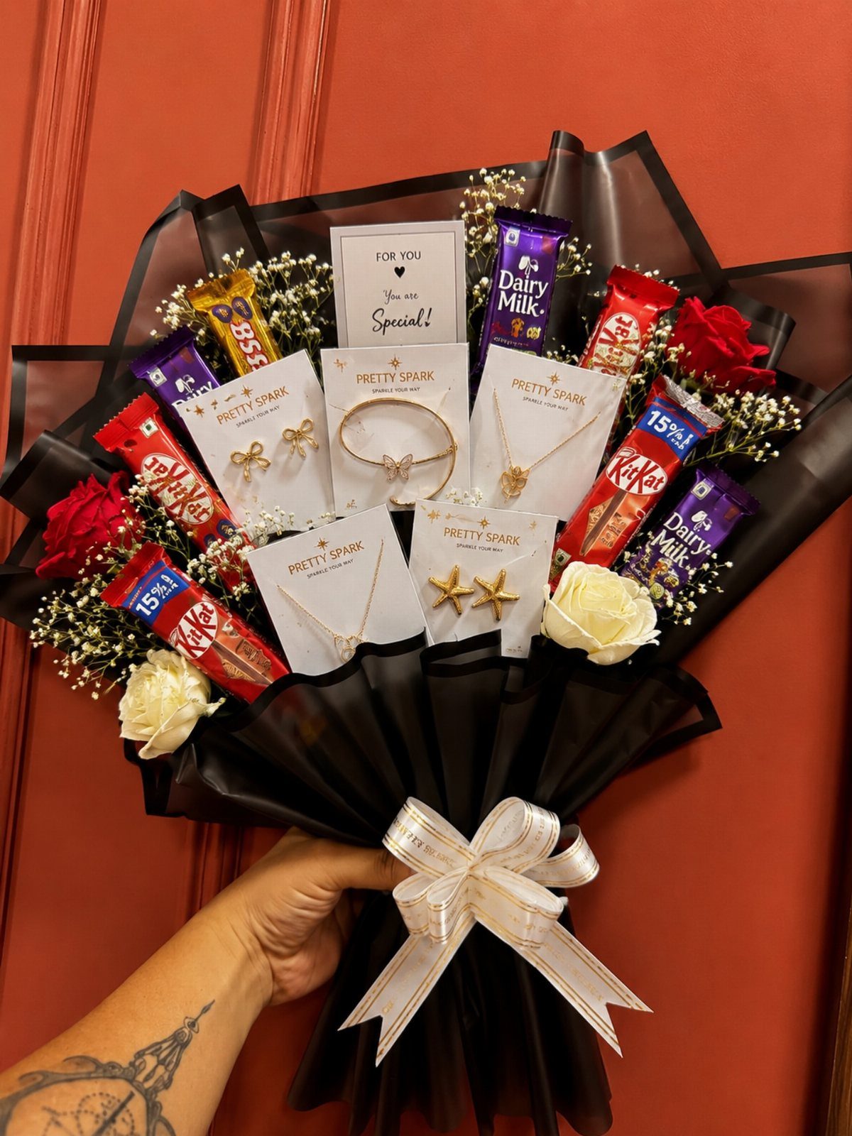 Bouquet of chocolates, gold jewelry, and roses wrapped in black paper with white bow.