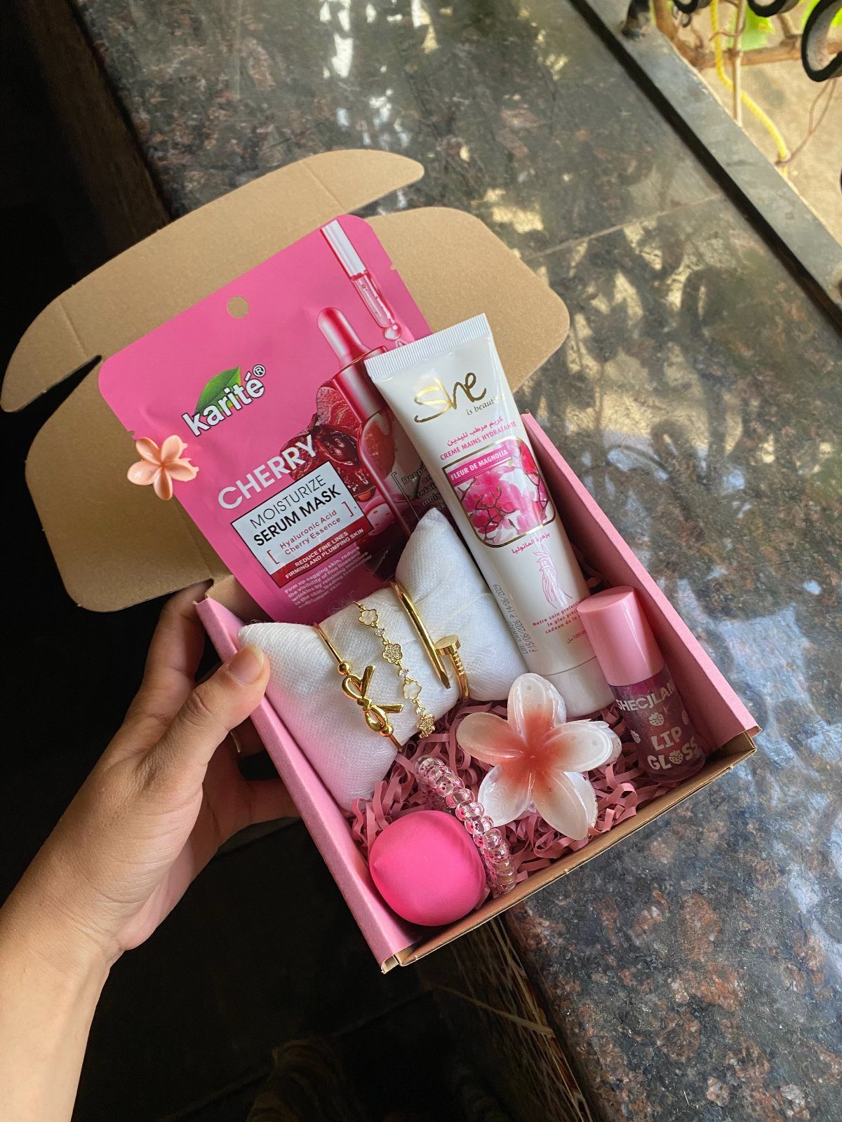 Pink gift box filled with skincare, jewelry, and flowers held by a hand.