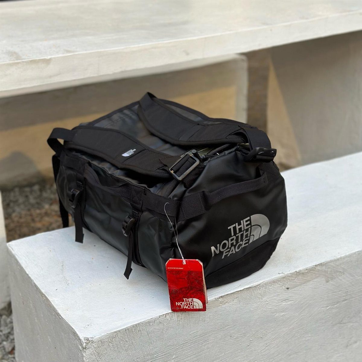 Black The North Face duffle bag with red tag on white concrete ledge.
