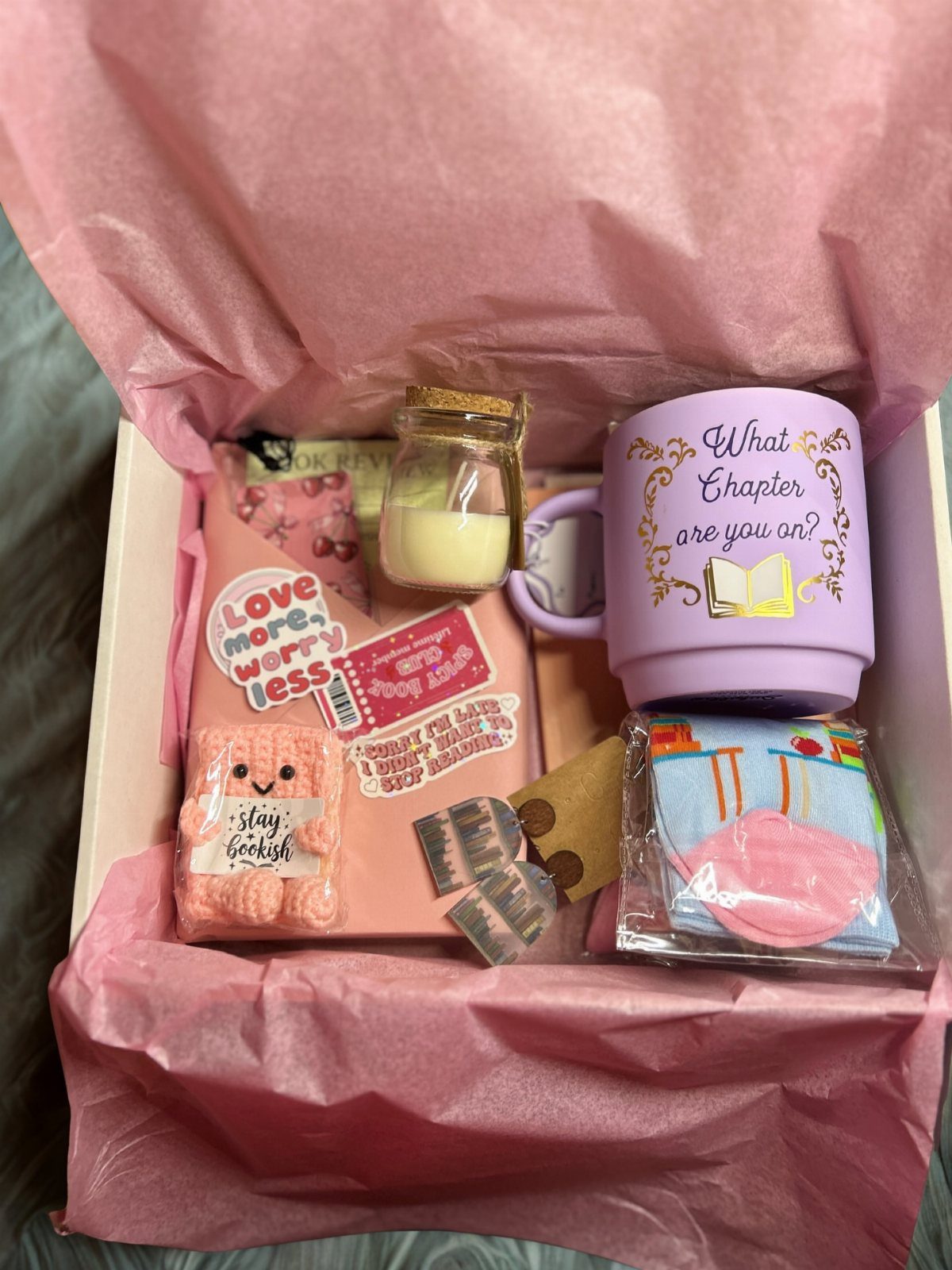 Gift box with a purple book-themed mug, candle, notebook, and cozy accessories.