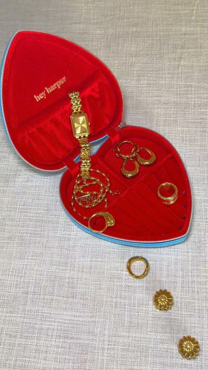 Heart-shaped jewelry box with red velvet interior holding gold jewelry on grey fabric.