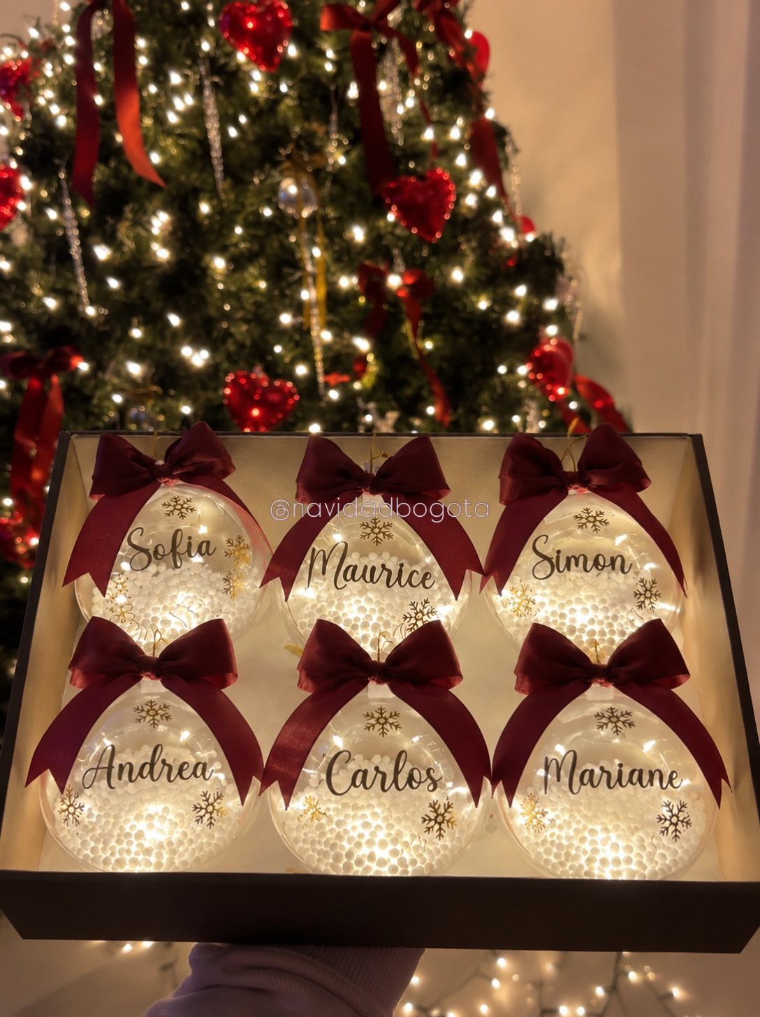 Six personalized light-up Christmas ornaments with names and burgundy bows in a box.