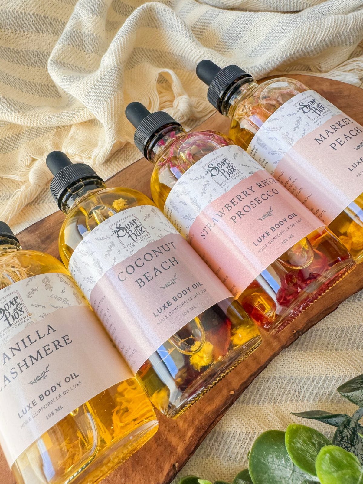 Four bottles of scented luxe body oil on a wooden board with fabric.