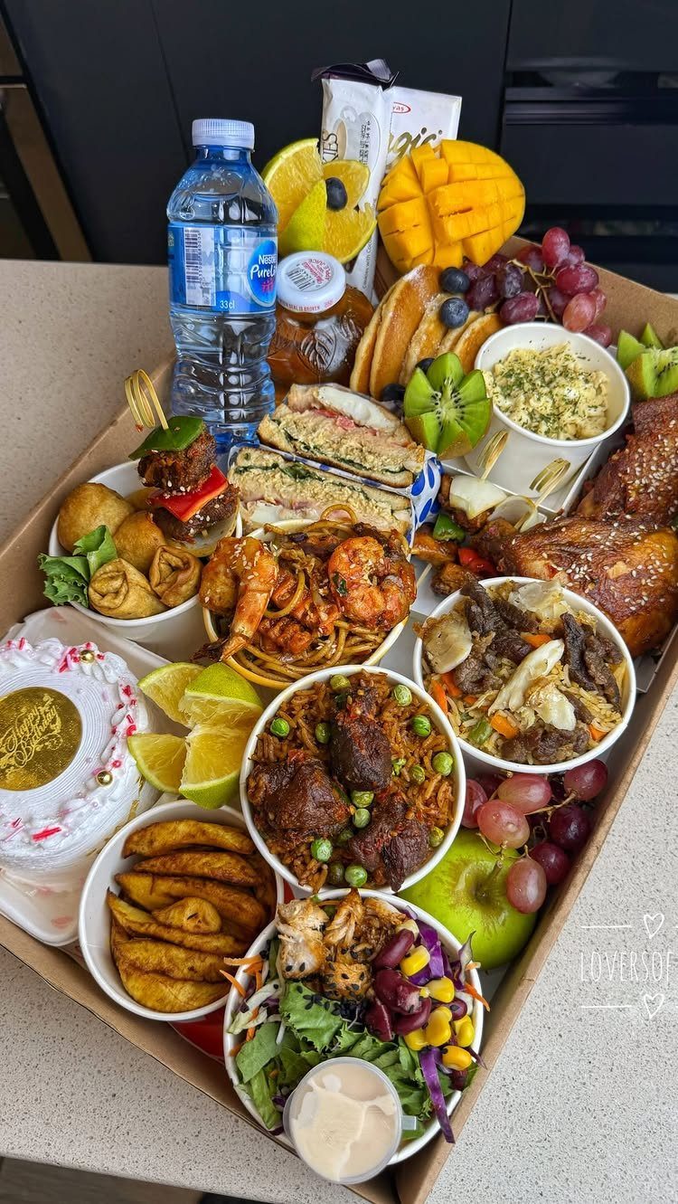 Cardboard gift box filled with diverse savory foods, fresh fruits, and a small cake.