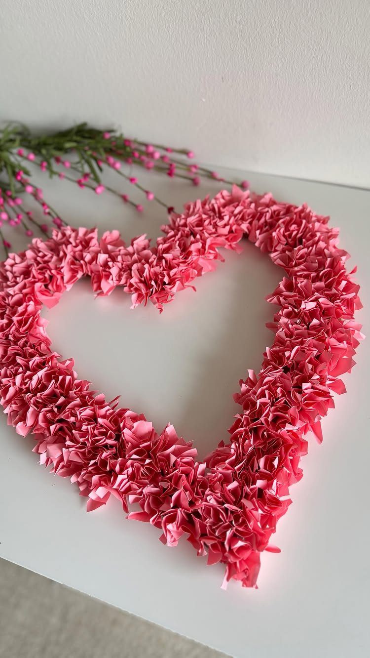 Pink paper heart wreath resting on a white surface with green foliage.