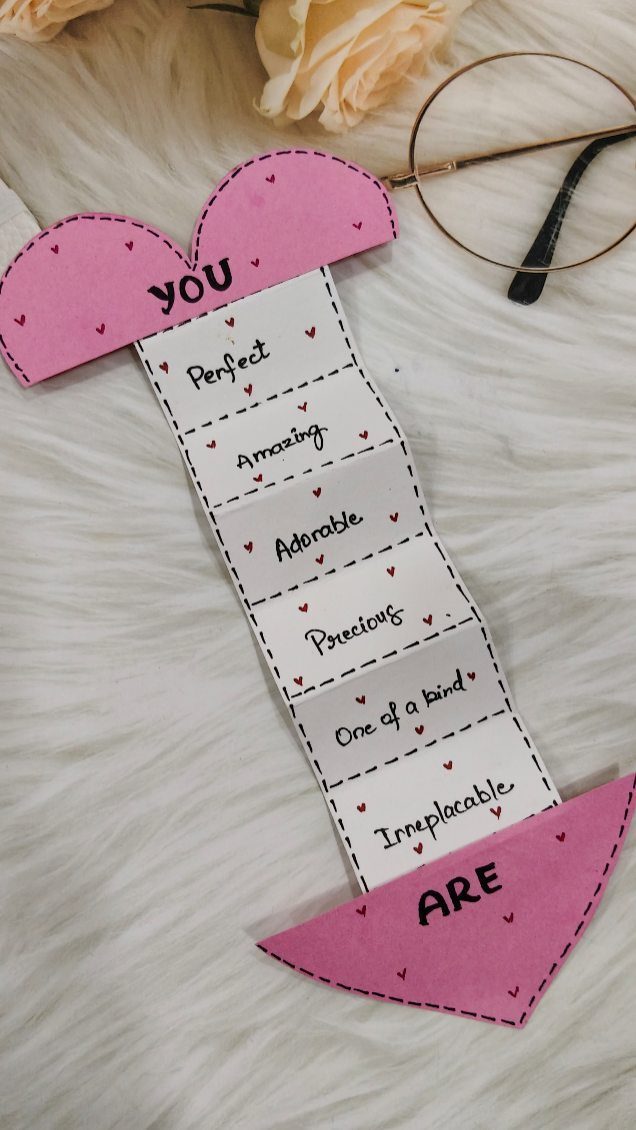 Handmade pink and white paper heart craft with handwritten compliments on a white rug.