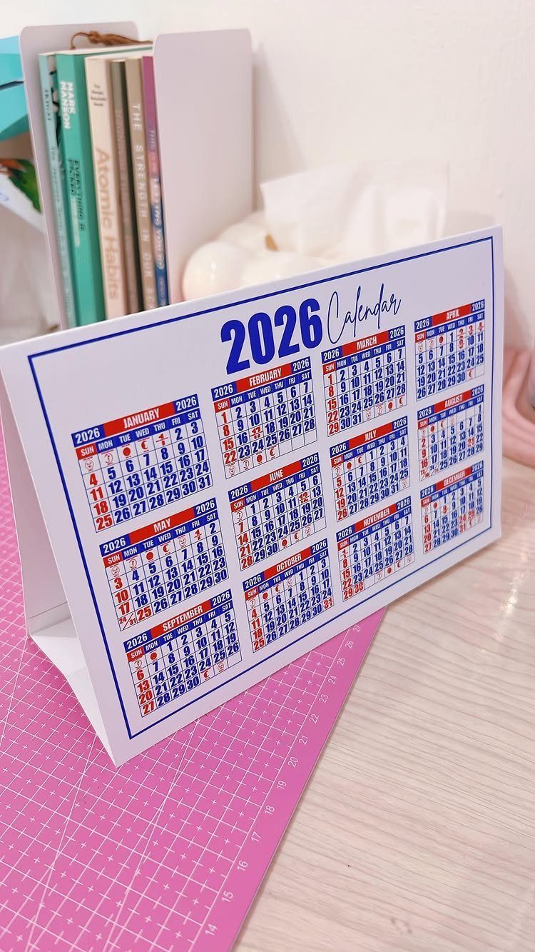 White 2026 desk calendar standing on a pink cutting mat next to books.