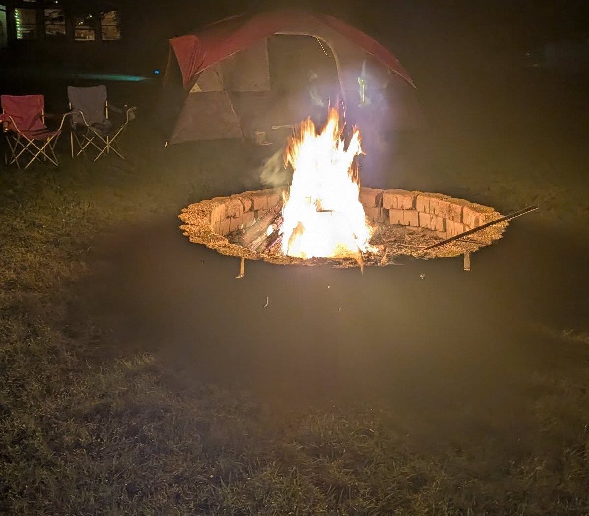 Campfire in a stone ring with a red tent and chairs at night.