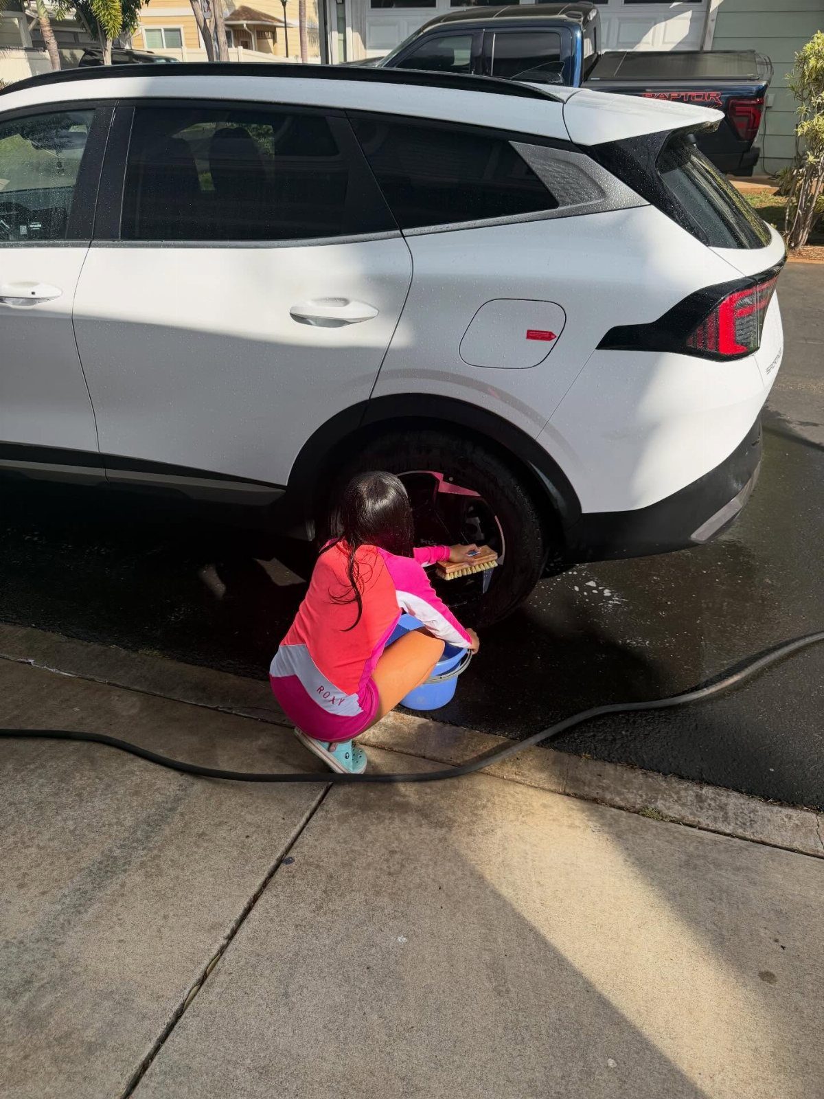 Child in pink outfit scrubbing the wheel of a white SUV on a driveway