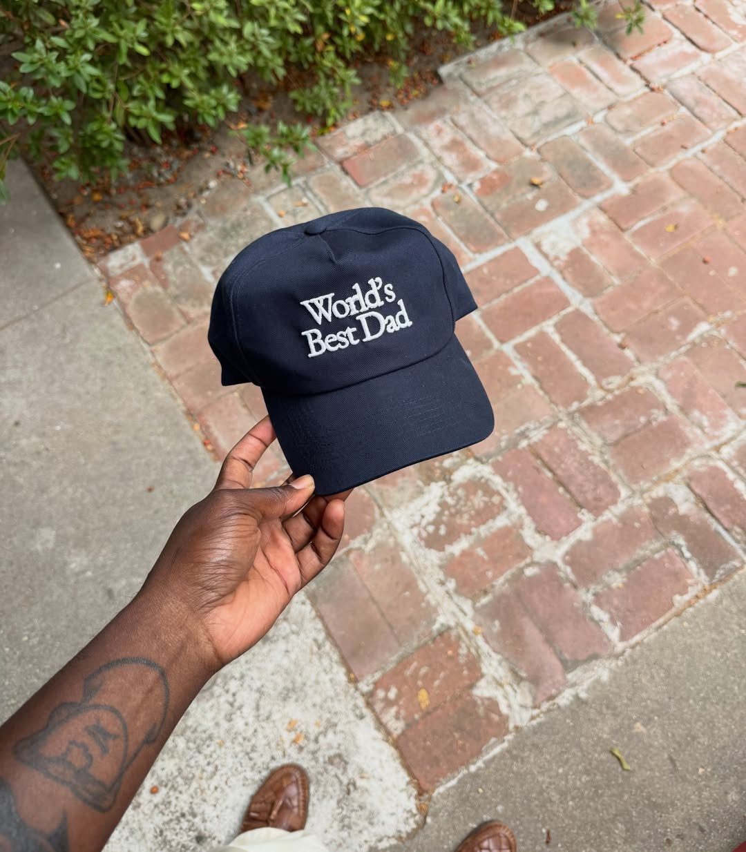 Dark blue baseball cap with World's Best Dad embroidery held over brick walkway