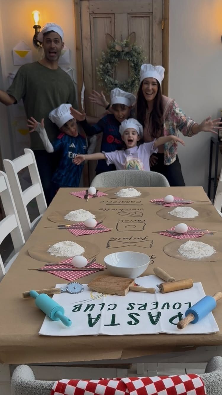 Family wearing chef hats posing behind a table set for homemade pasta making.