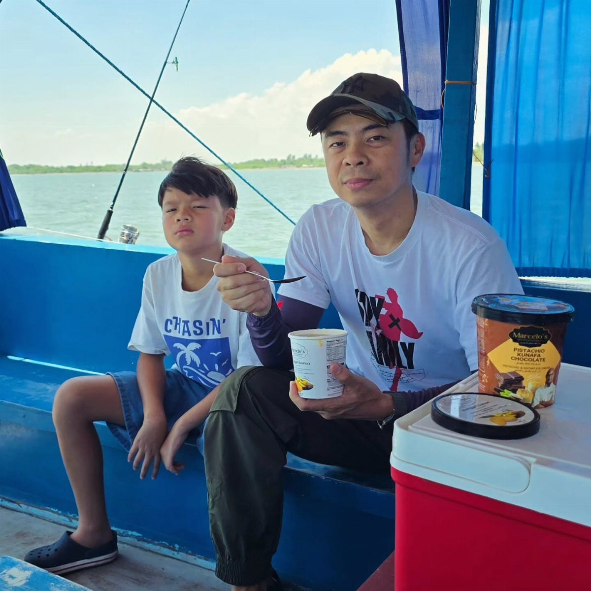 Man and young boy sitting on a blue boat with fishing rods nearby.