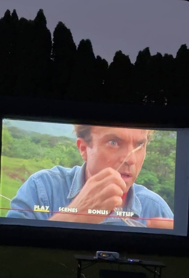 Outdoor movie screen showing a film scene with a projector on a table.
