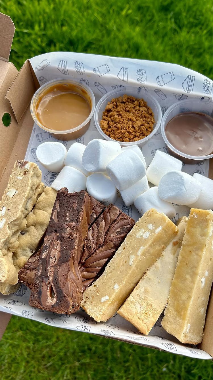 Assorted fudge, marshmallows, and dips in a cardboard snack box on green grass.
