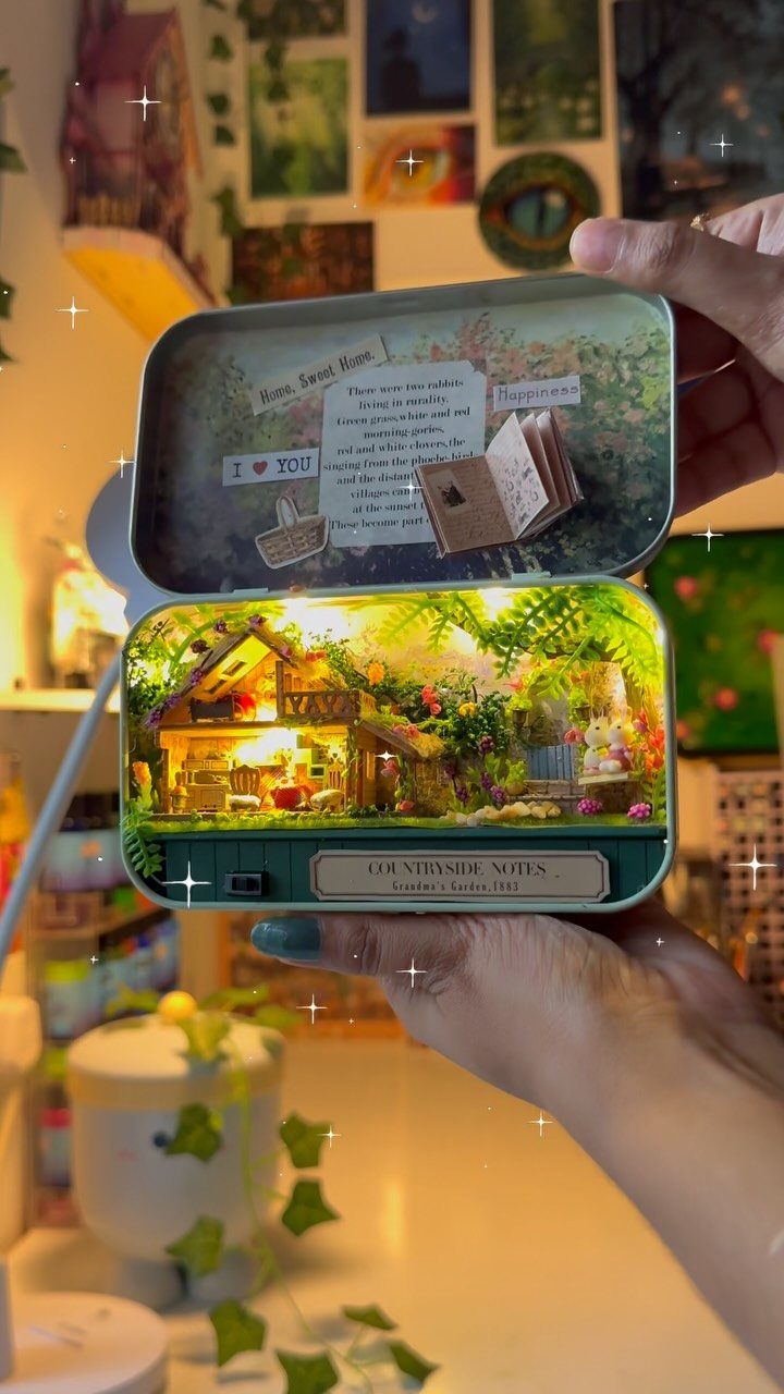 Miniature illuminated cottage diorama inside a metal tin held by two hands.