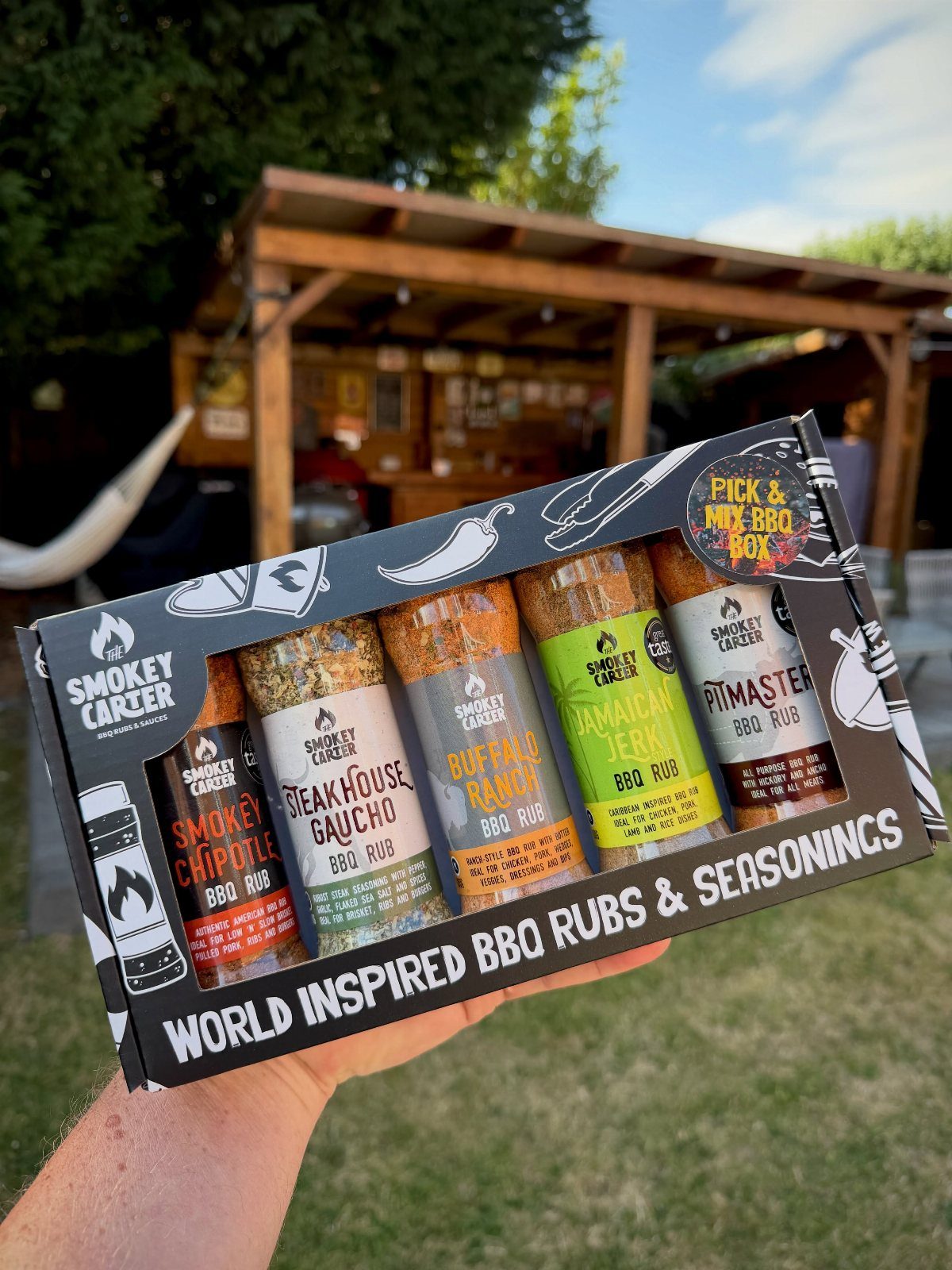 Hand holding a gift box containing five jars of assorted BBQ rub seasonings.