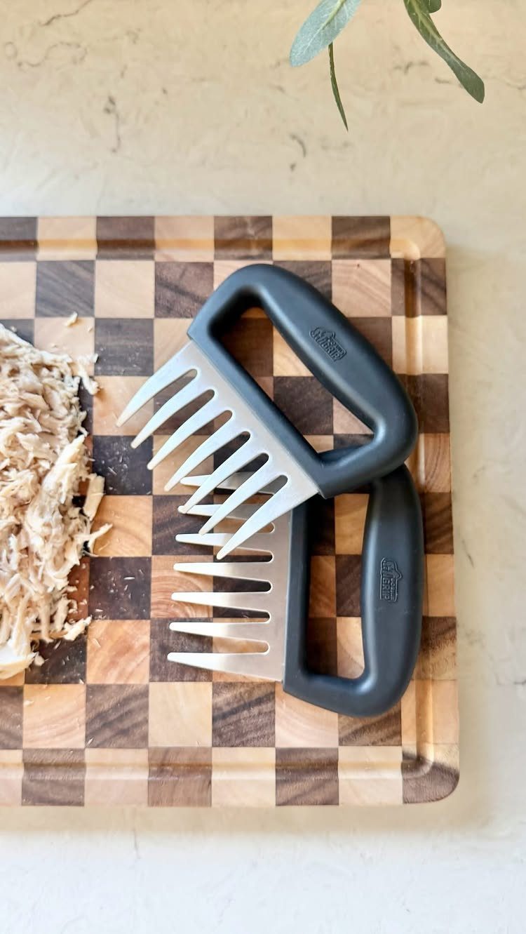 Meat shredder claws with black handles on a checkered wooden cutting board.