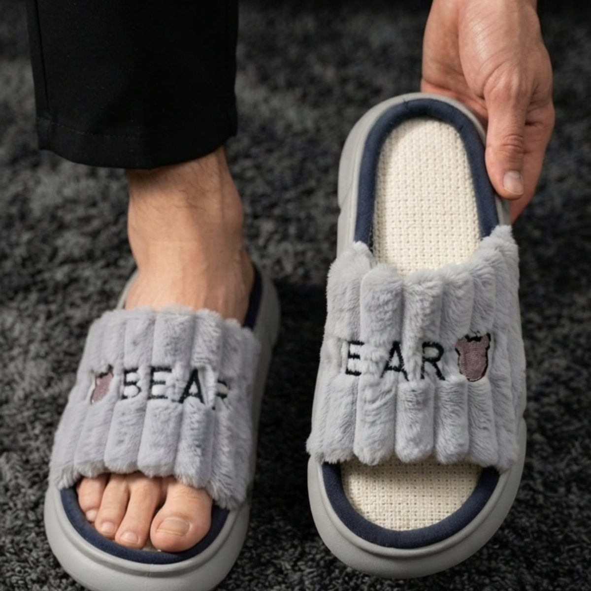 Grey fuzzy BEAR slippers with thick rubber soles on a dark grey carpet.