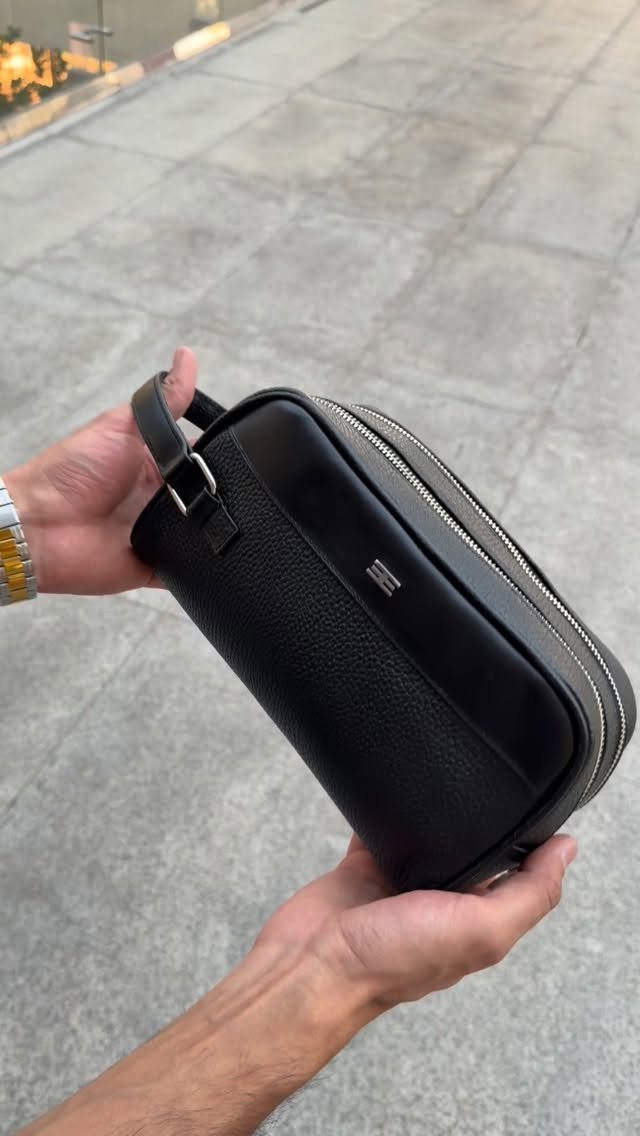Black pebbled leather crossbody bag held by hands over a grey concrete surface.