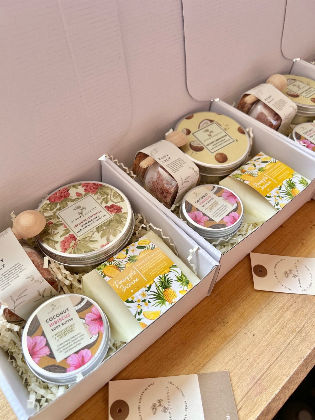 Three white gift boxes filled with body butter, soaps, and skincare products on wood.
