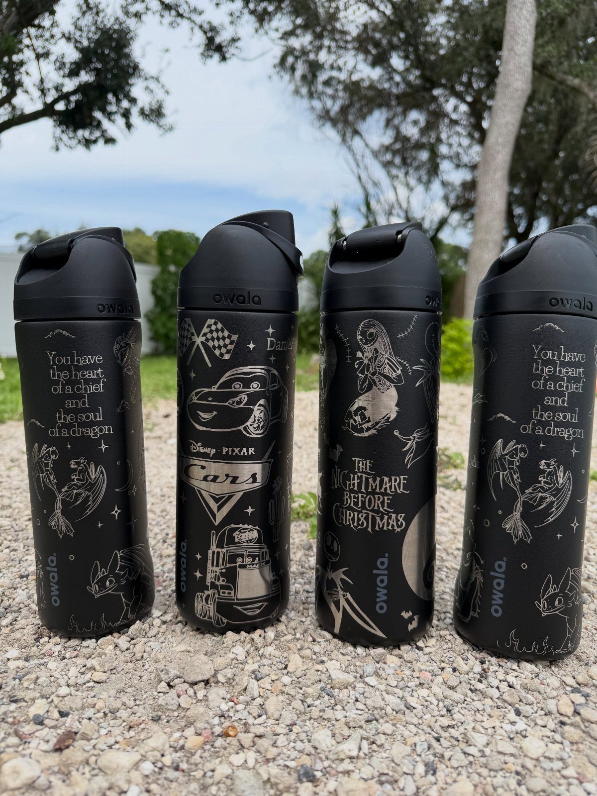 Four black personalized tumblers with custom graphics standing on a gravel surface.