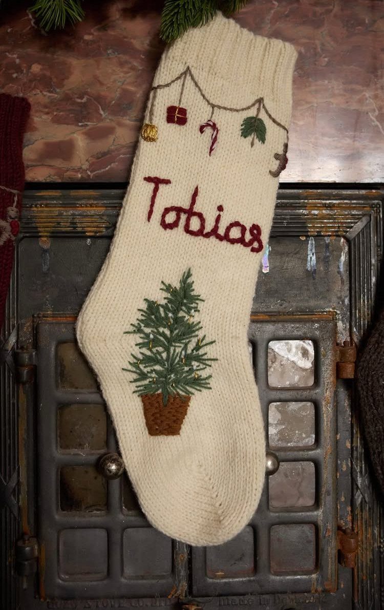 Cream knitted Christmas stocking with name Tobias and tree hanging by a fireplace