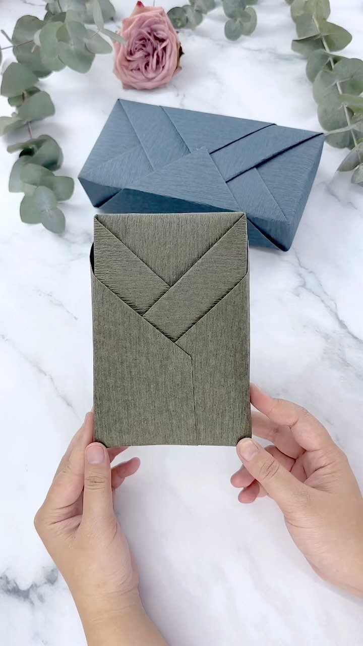 Hands holding an olive green wrapped gift box with geometric folds on marble.