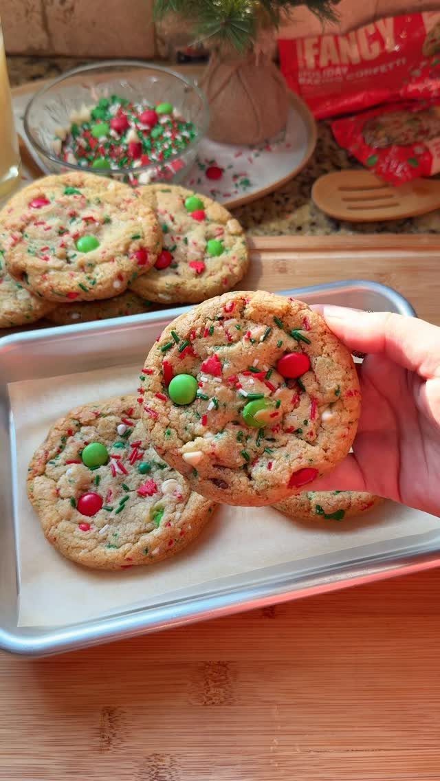 Hand holding a colorful cookie with red and green sprinkles on a baking sheet