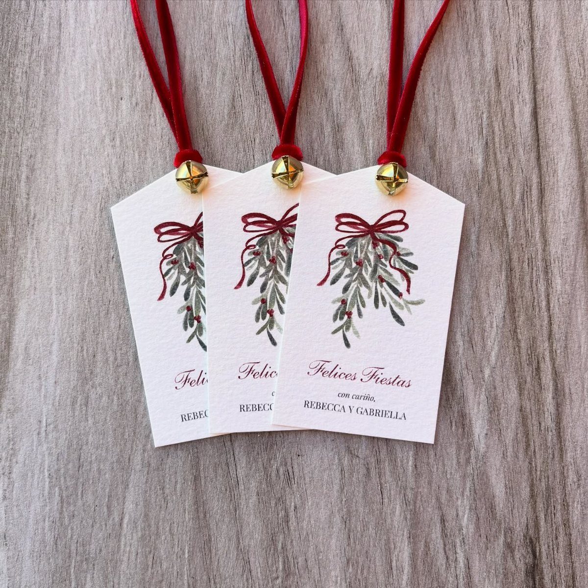 Three white Christmas gift tags with red ribbons, gold bells, and botanical illustrations.