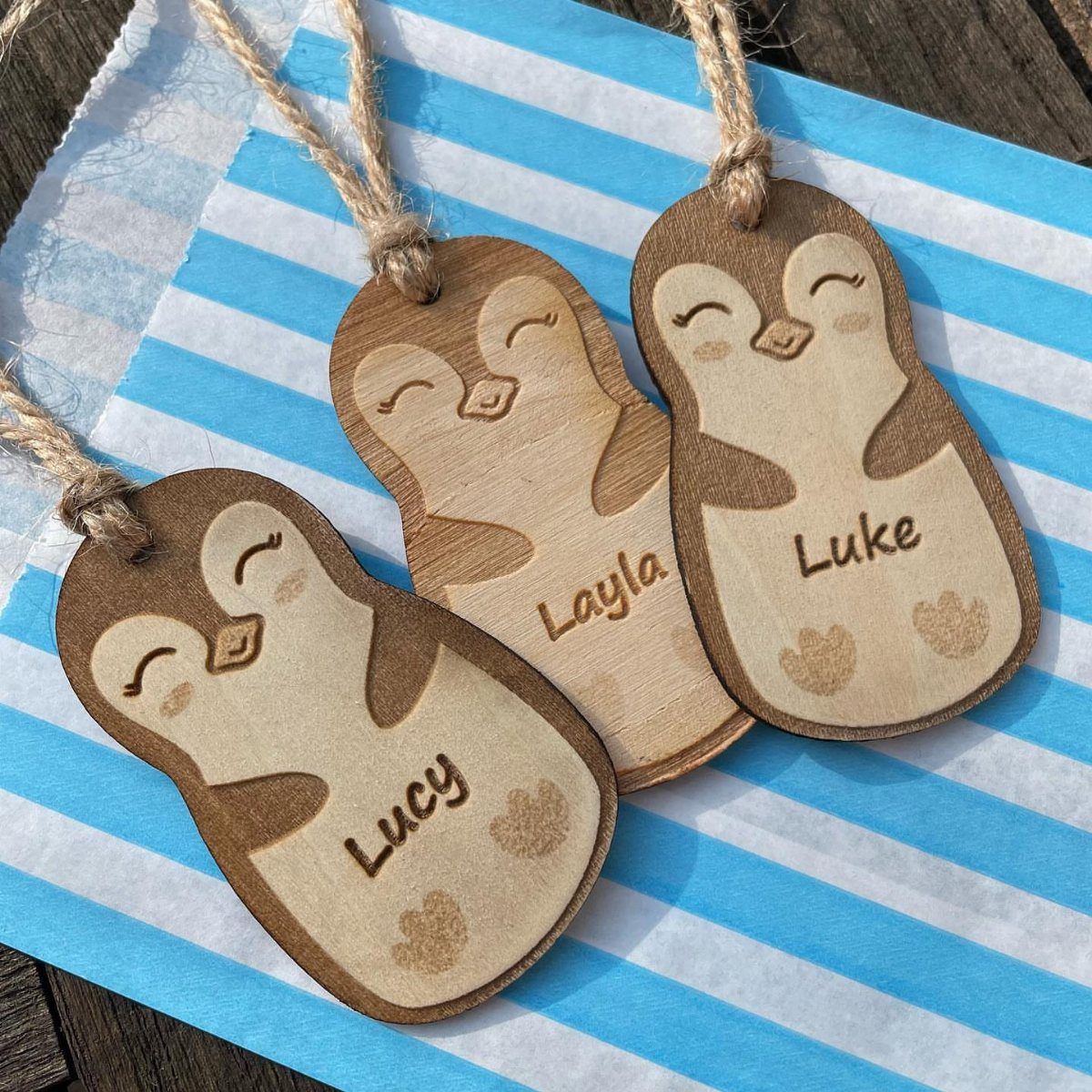 Wooden penguin gift tags with names engraved, resting on blue striped paper.