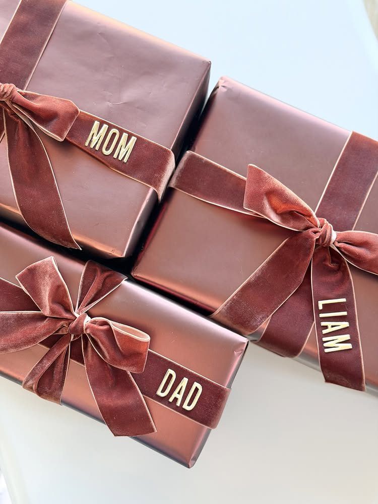 Three rose-gold wrapped gifts with velvet ribbons and gold name tags on white.
