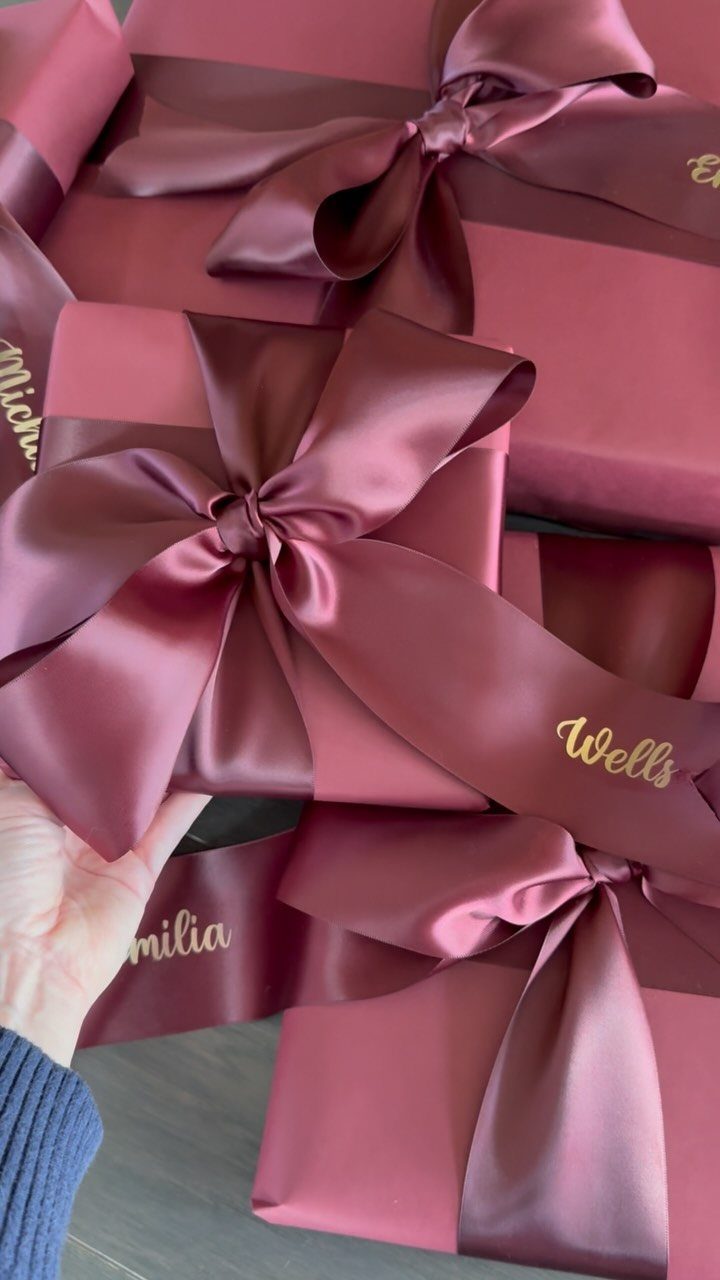 Mauve-pink wrapped gift boxes with large satin bows and gold cursive names.