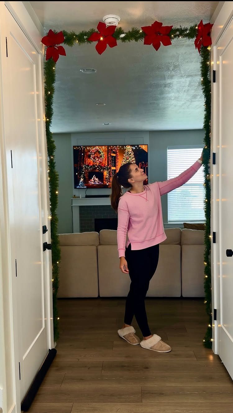Woman decorating a doorway with green garland, red poinsettias, and fairy lights.