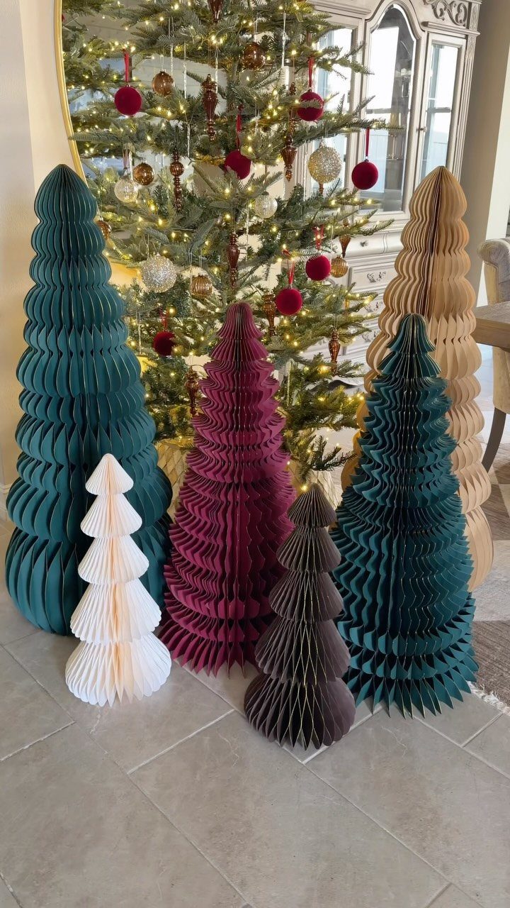 Honeycomb paper decorative trees in various colors standing before a decorated Christmas tree.