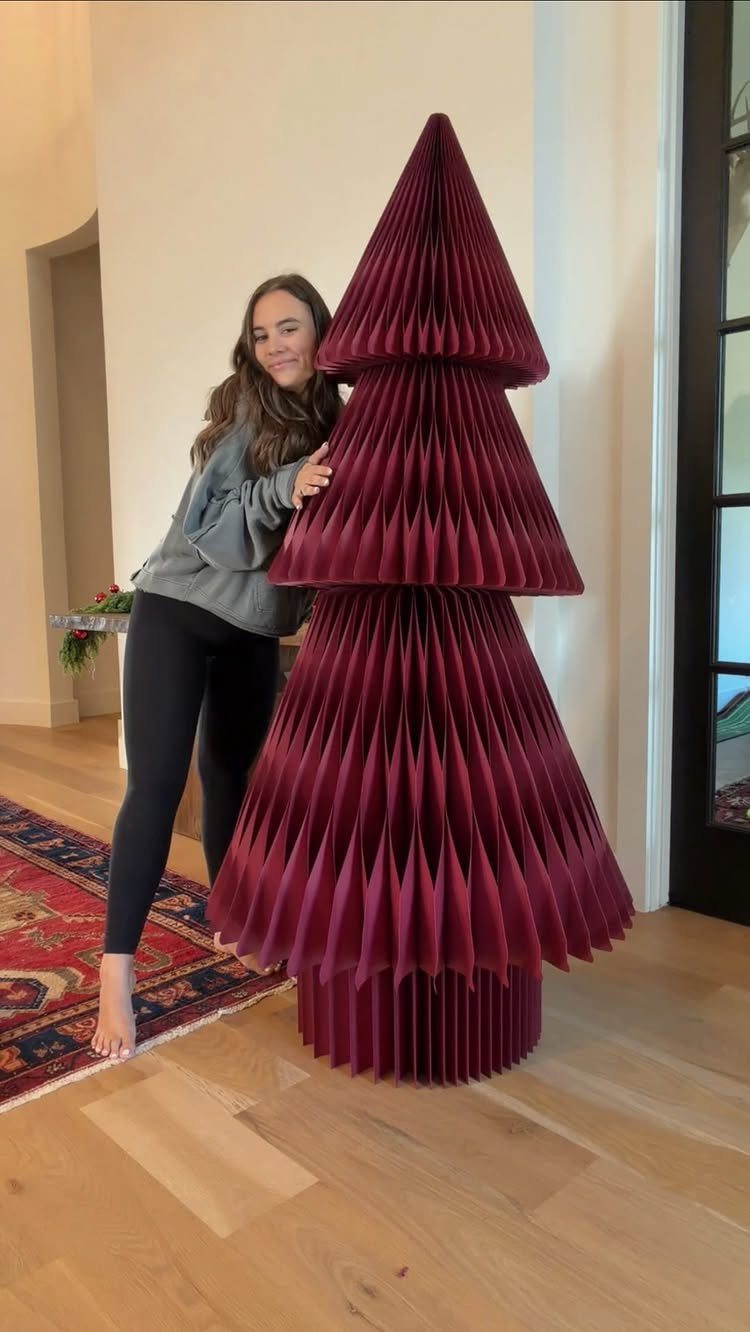 Woman standing beside a large burgundy accordion-folded paper Christmas tree decoration.