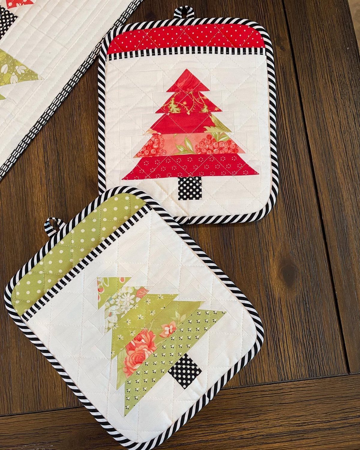 Two quilted Christmas tree potholders in red and green on a wooden table.