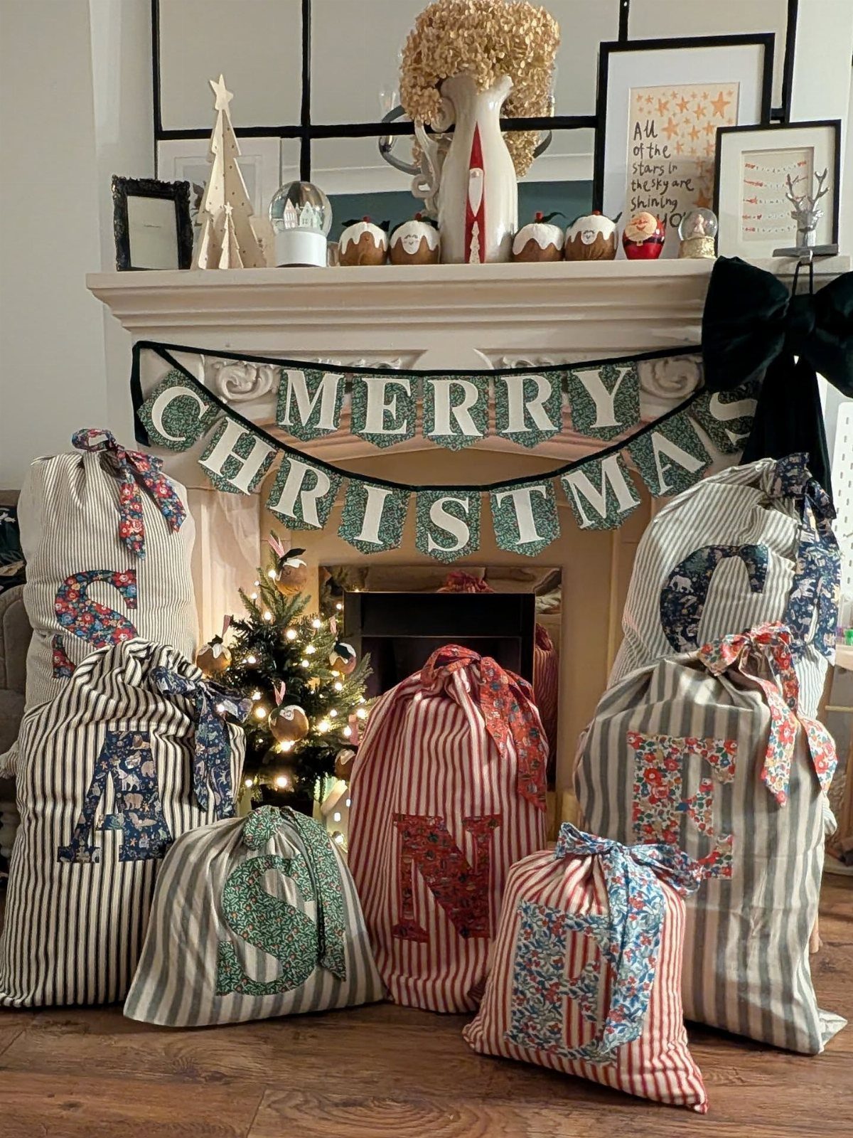 Large striped fabric Santa sacks with floral letters arranged by a fireplace.