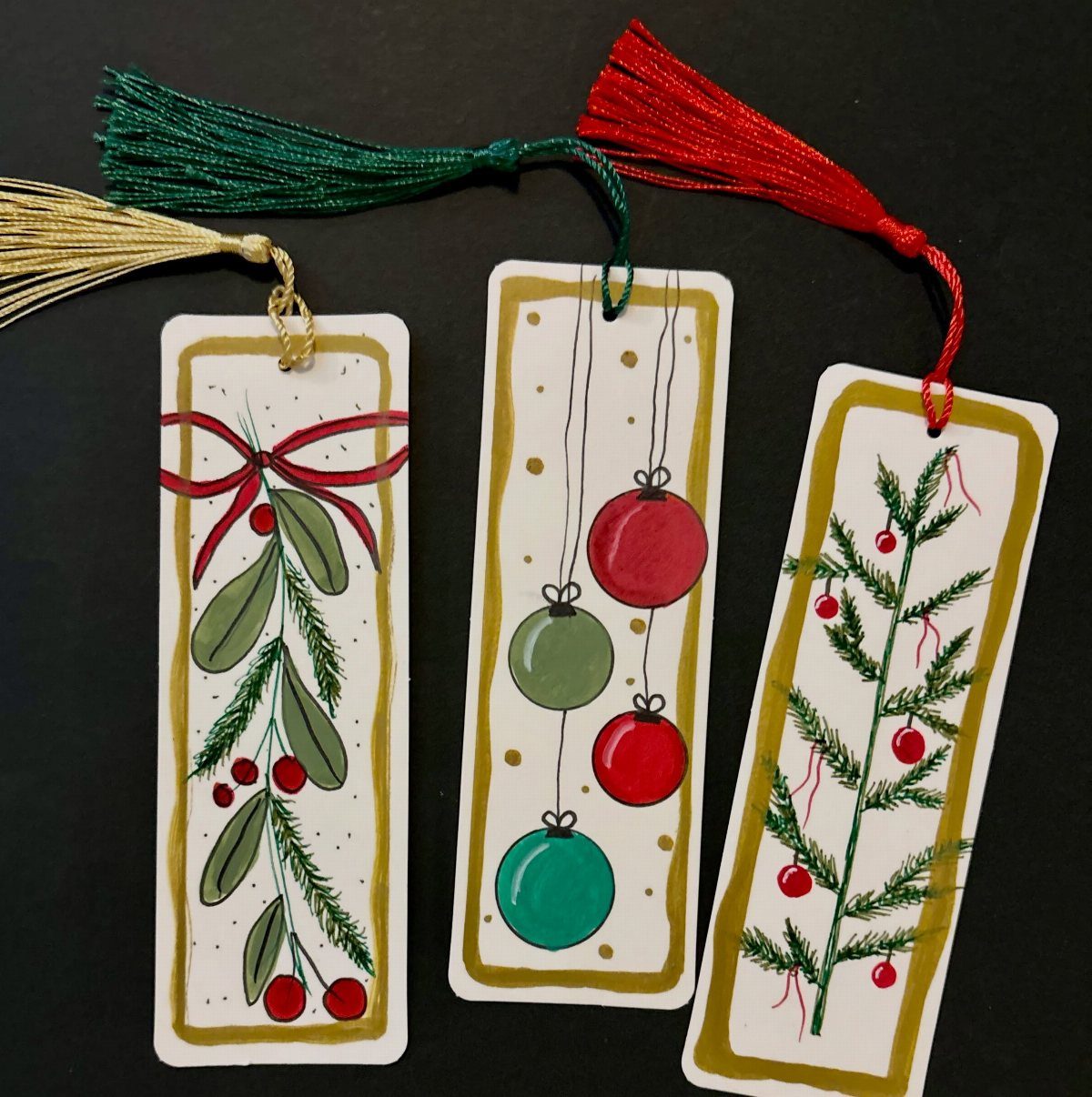 Three handmade Christmas bookmarks with tassels and festive drawings on a black surface.