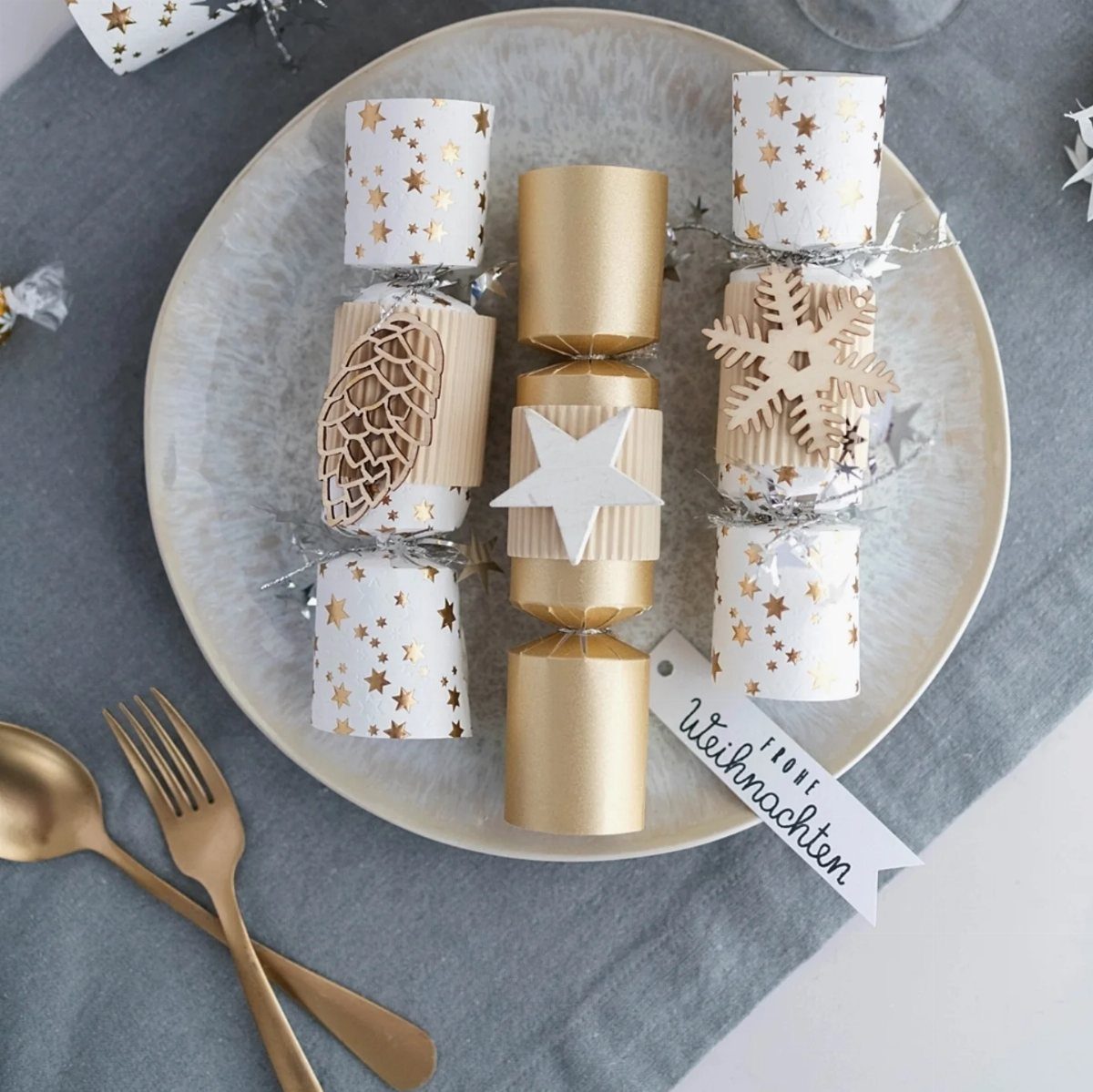 Christmas crackers decorated with wooden shapes on a plate with gold cutlery.