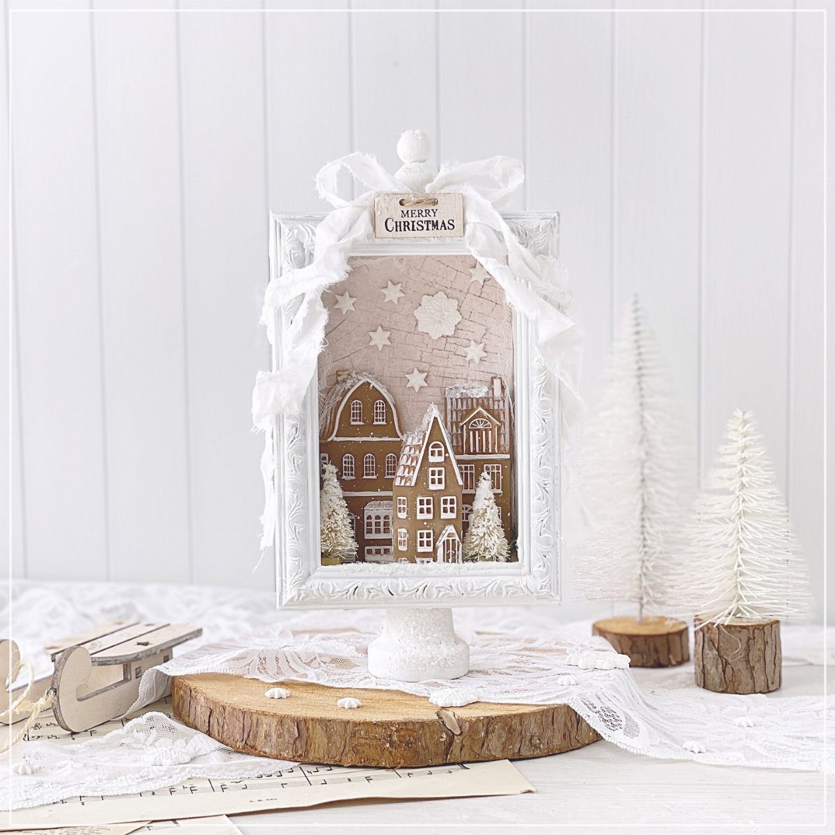 Miniature gingerbread village inside a white decorative frame on a rustic wood slice