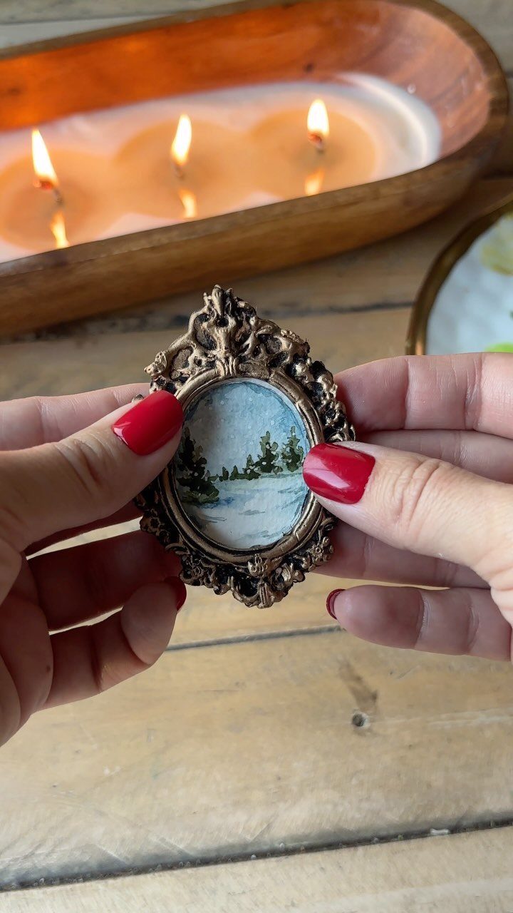 Small ornate gold frame with winter landscape painting held by red-nailed hands.