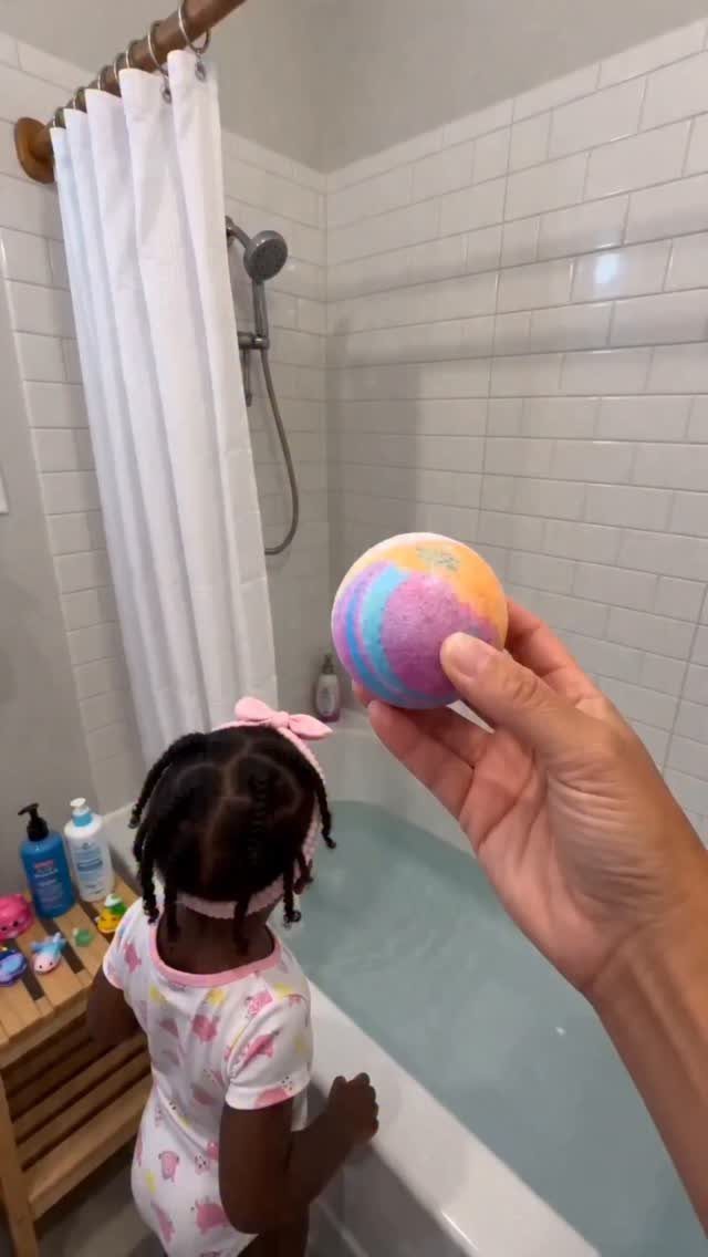 Colorful bath bomb held in hand with a child standing by a bathtub.