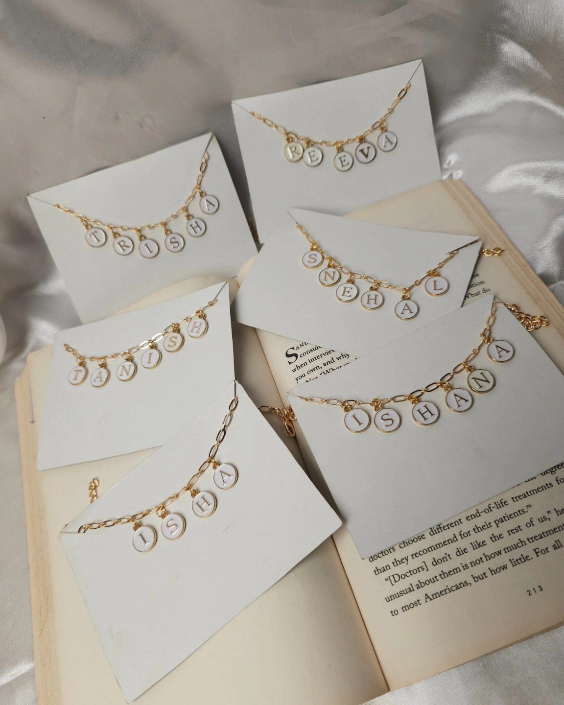 Gold chain bracelets with white letter charms spelling names on white display cards.