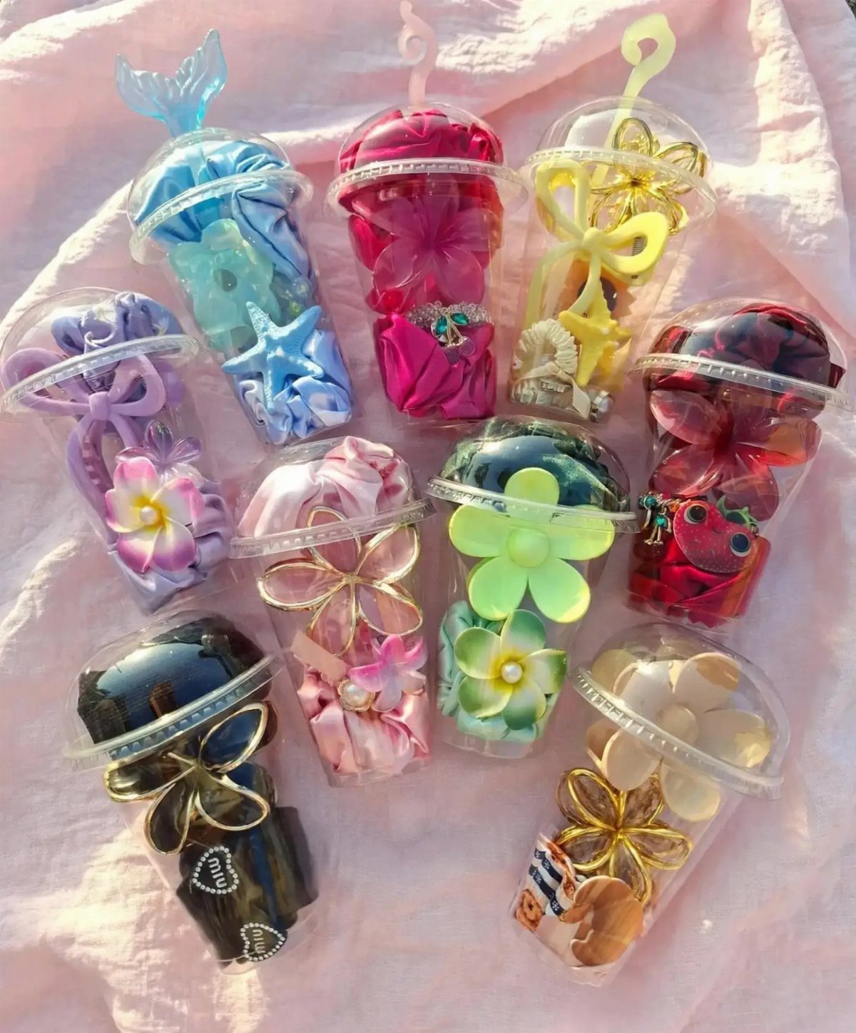 Colorful hair clips and bows arranged in small clear plastic cups on pink fabric.