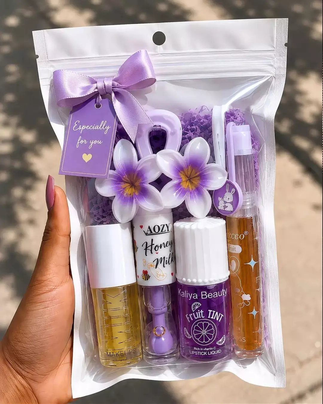 Purple themed beauty gift bag containing lip glosses, a pen, and decorative flowers.